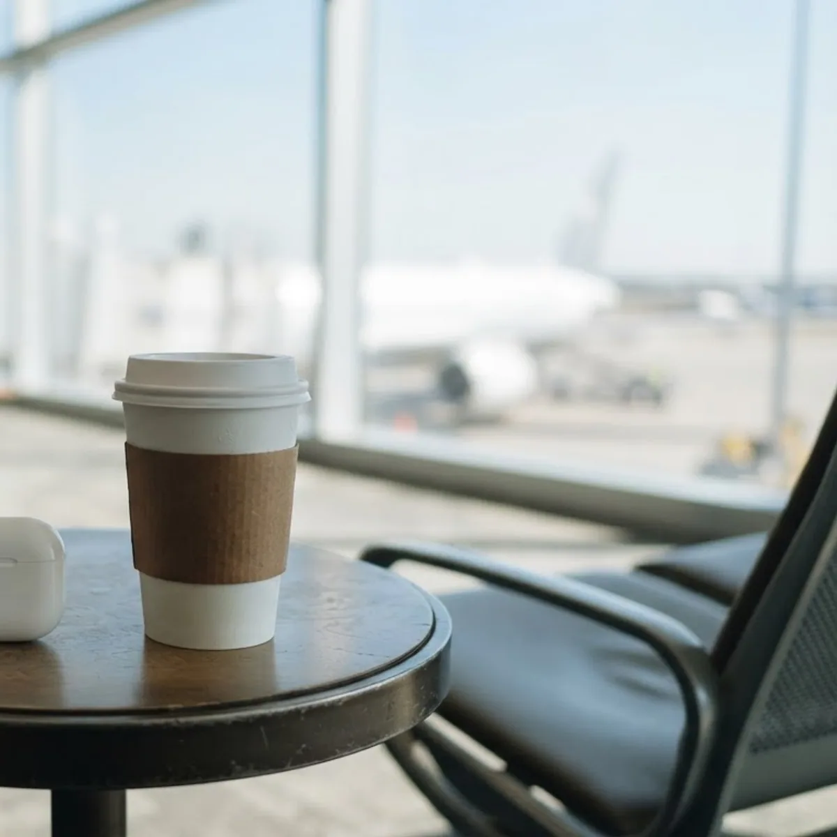 Closed white earbuds case beside a coffee cup in an airport lounge