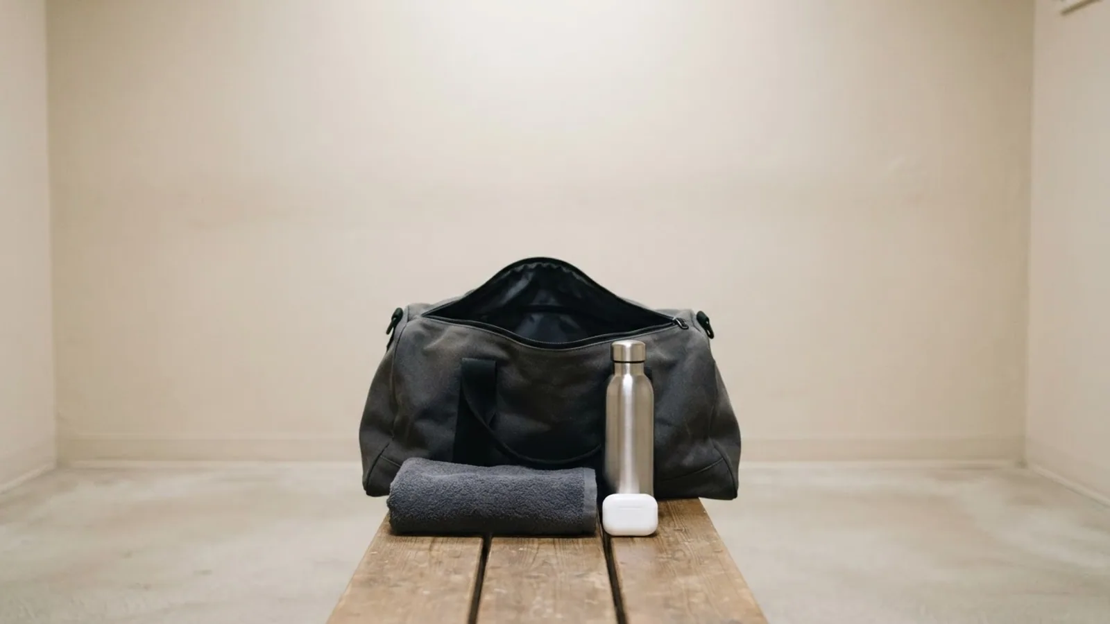 An open gym bag on a wooden bench with a folded towel, a steel water bottle and a closed white earbuds case