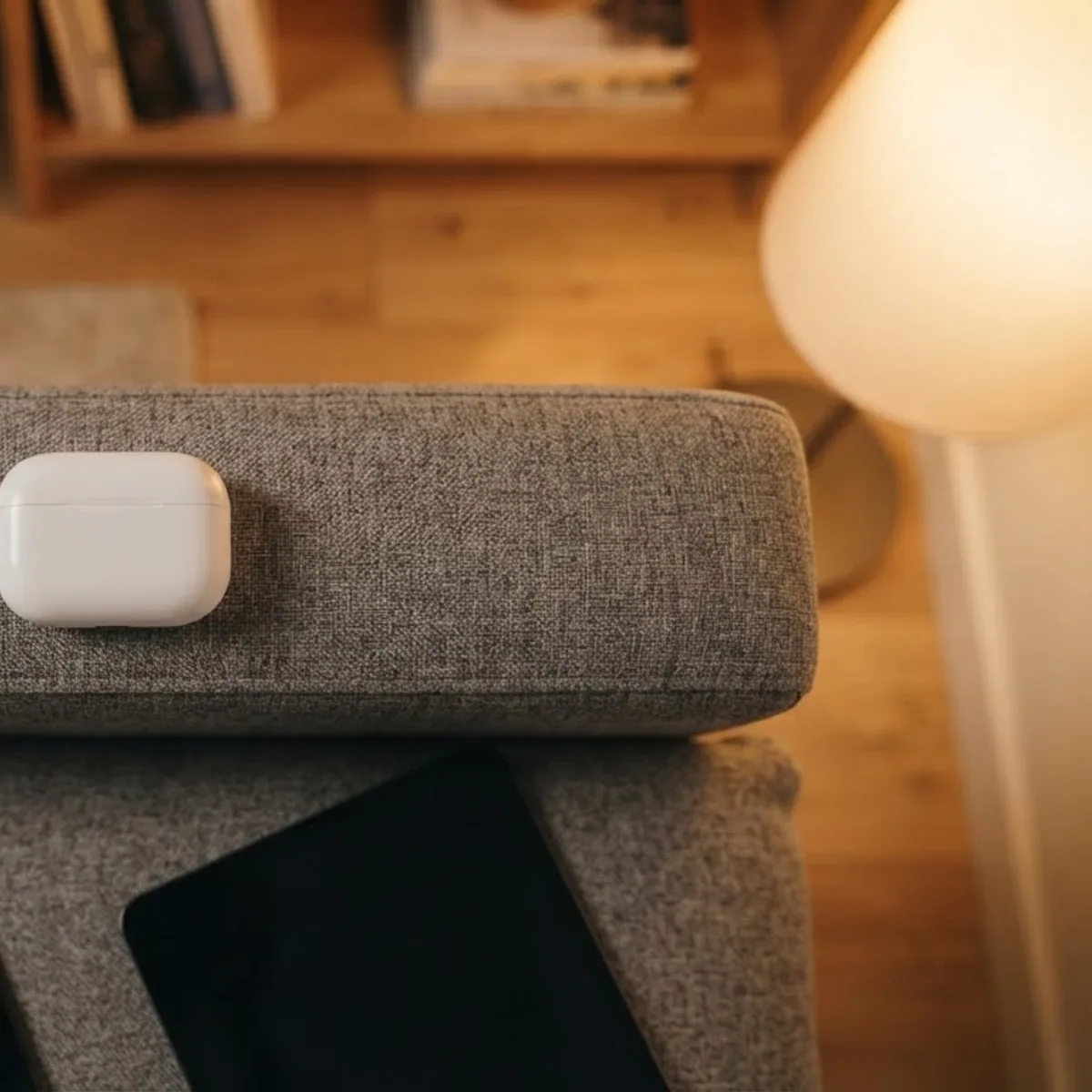 Top view of a closed white earbuds case on the arm of a grey sofa, with a phone and a tablet on the cushion below
