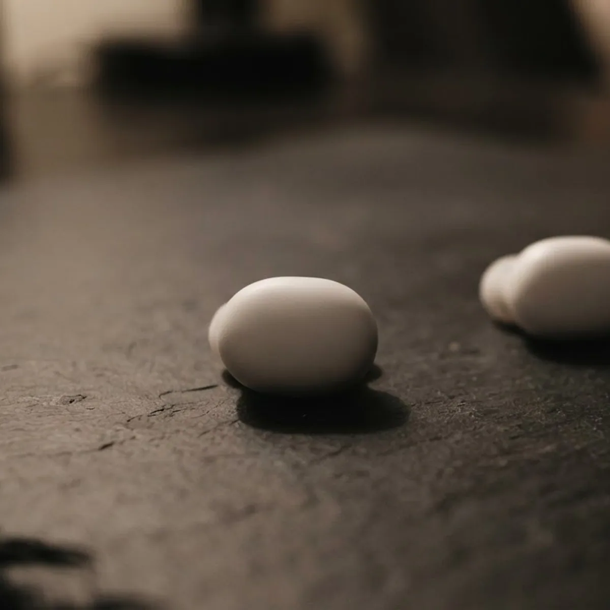 Two white wireless earbuds lying apart on a dark slate table in warm lamp light, one in sharp focus