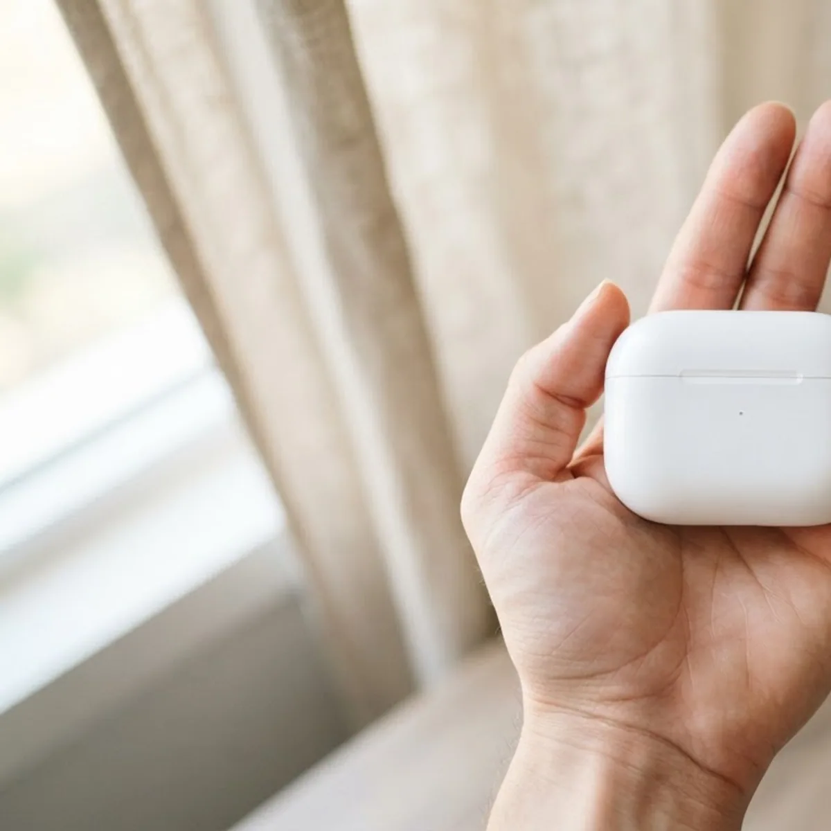 A closed white earbuds case resting in an open palm in front of linen curtains and a leafy plant