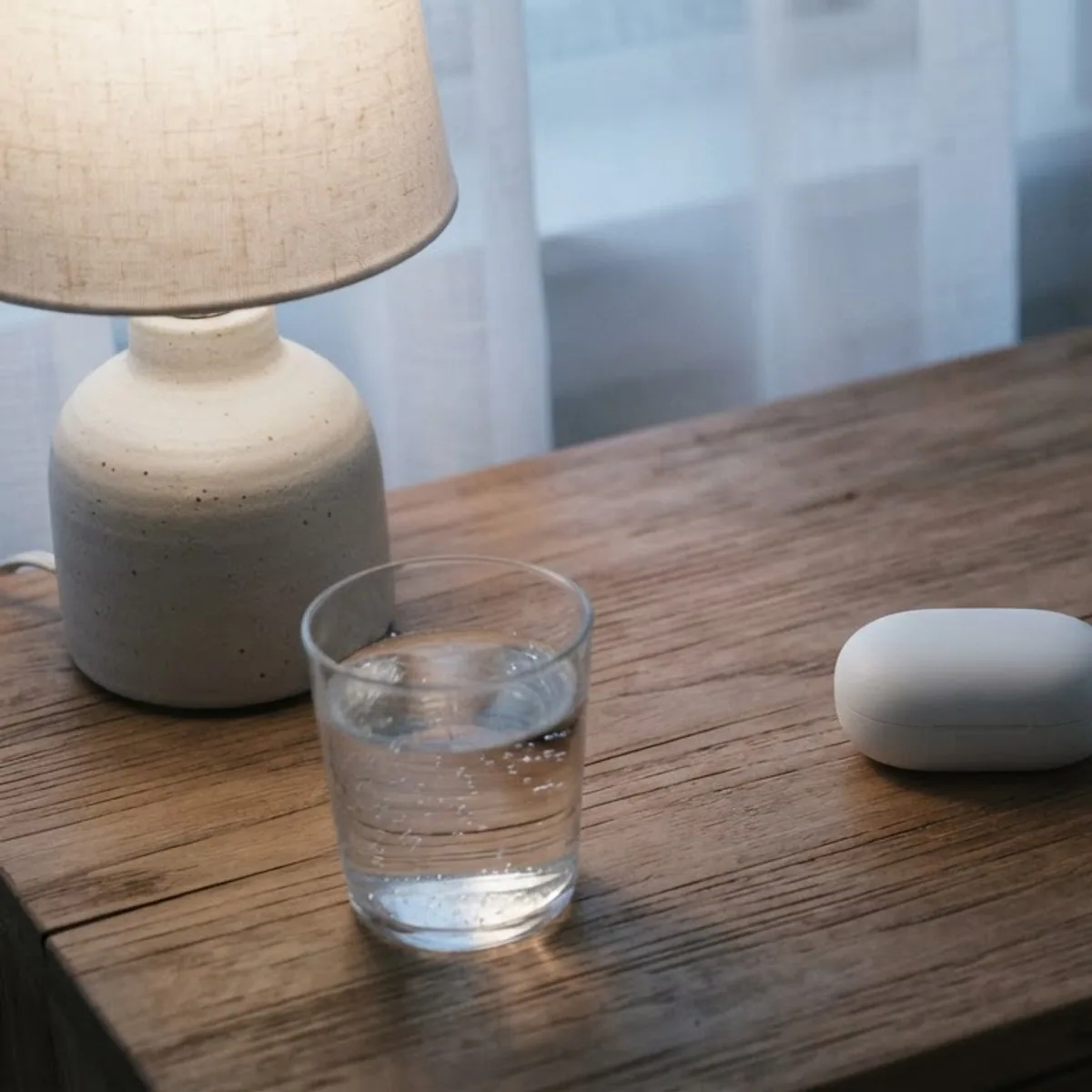 A closed white earbuds case next to a phone lying face down on a wooden bedside table with a glass of water and a lamp