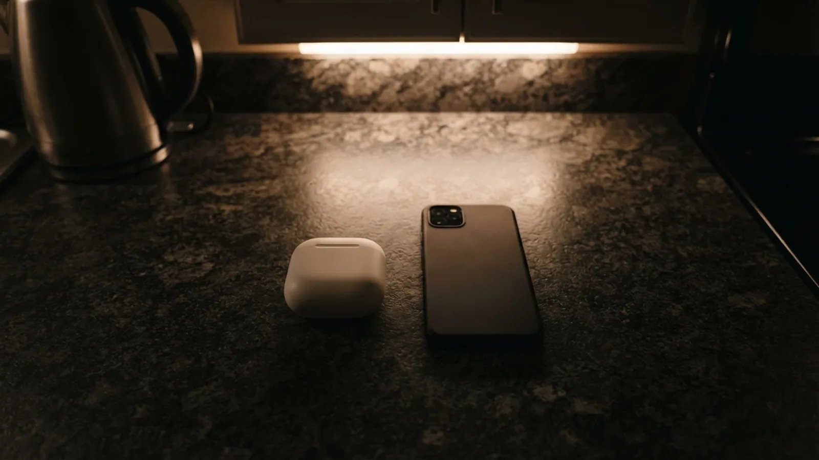 Top view of a closed white earbuds case and a phone lying face down on a dark stone kitchen counter at night under a warm cabinet light
