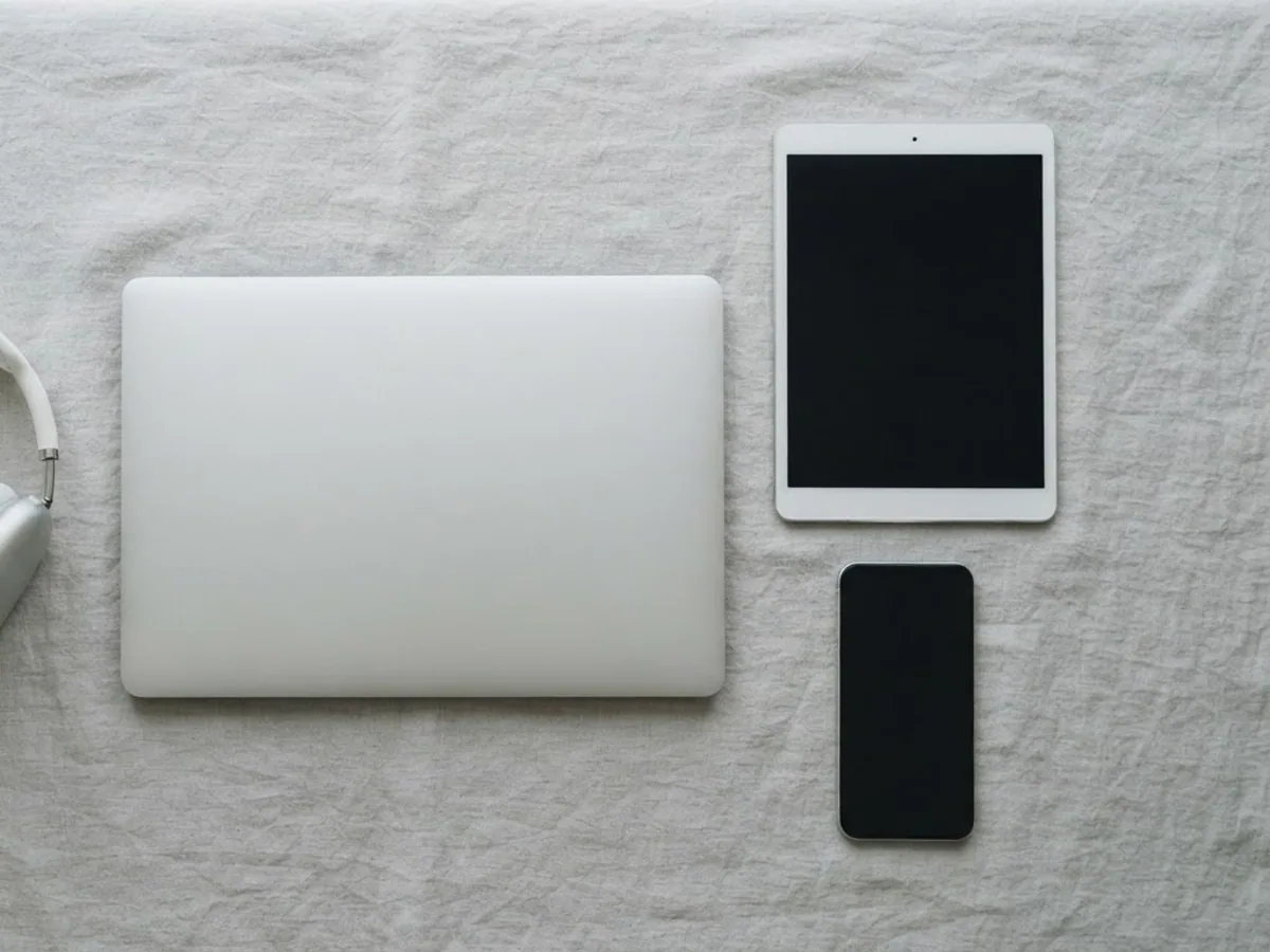 White over-ear headphones laid out next to a closed laptop, a tablet and a phone