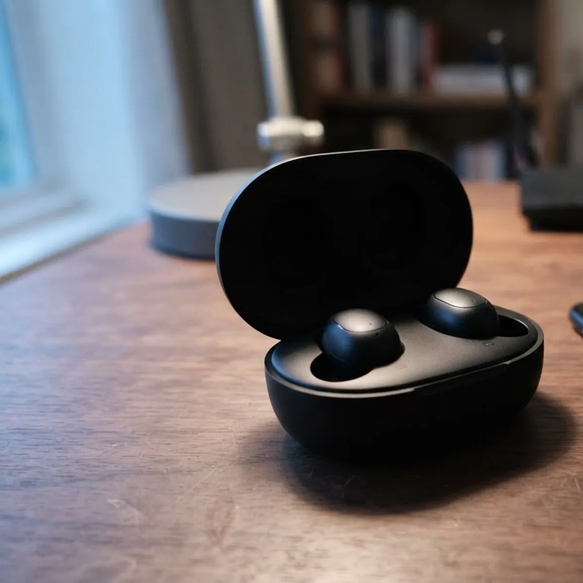 Black wireless earbuds in an open case beside a phone on a desk, with a router in the background