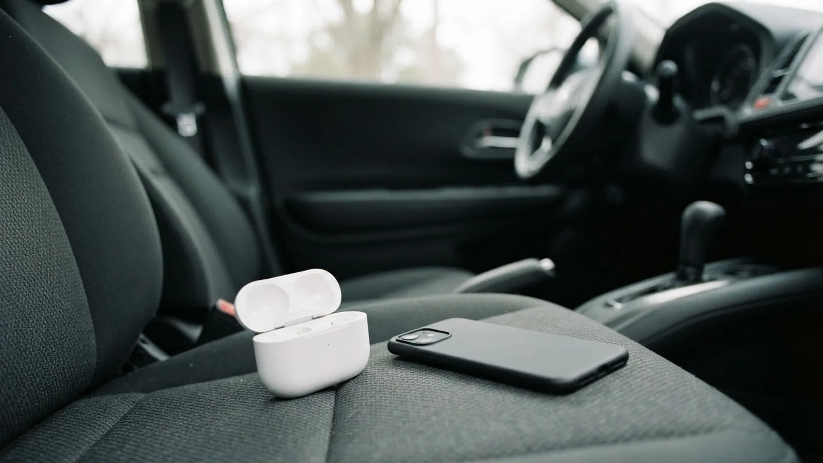 Open white earbuds case and a face-down phone on the passenger seat of a parked car