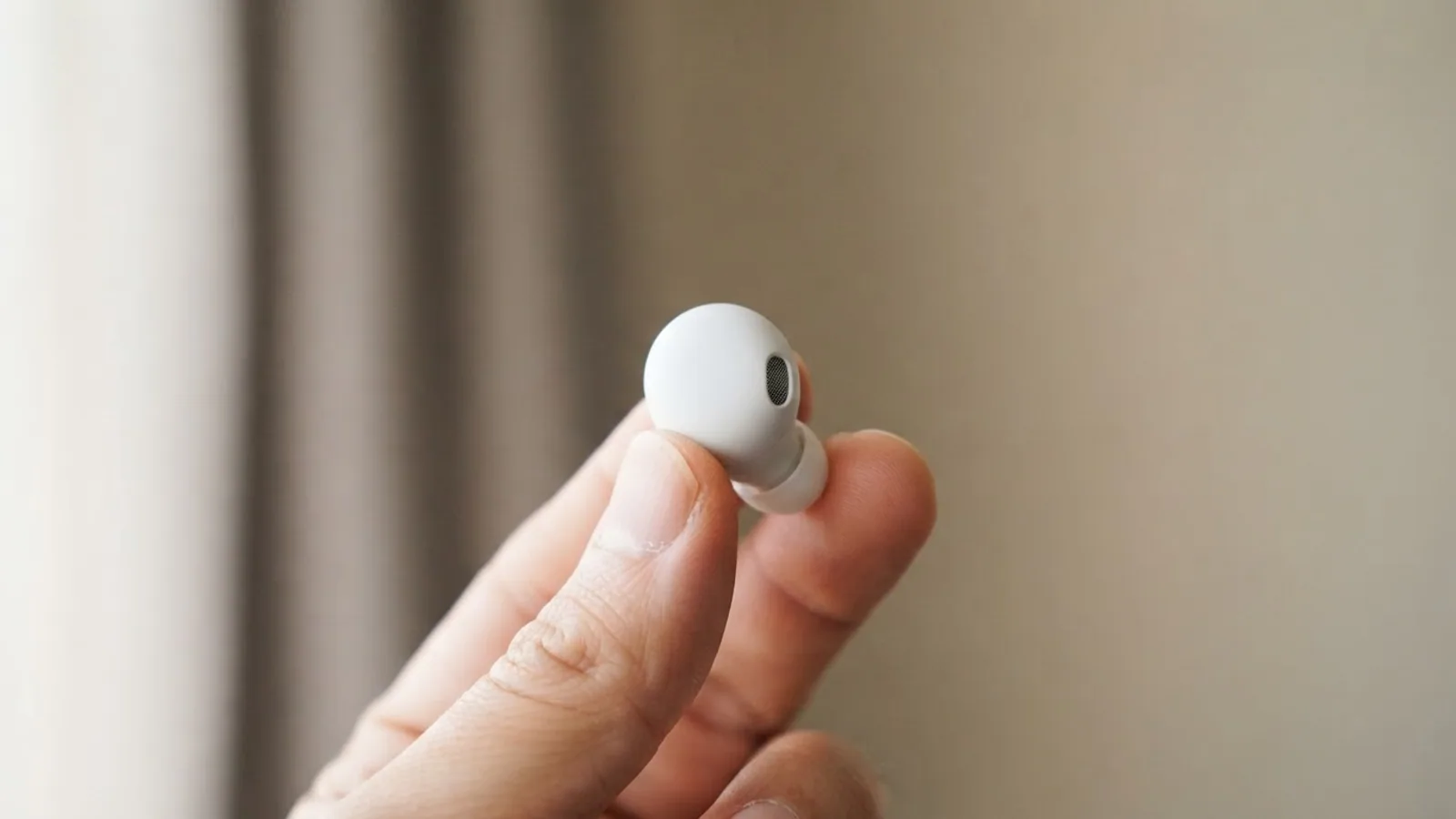Hand holding a single white earbud between thumb and finger in soft window light
