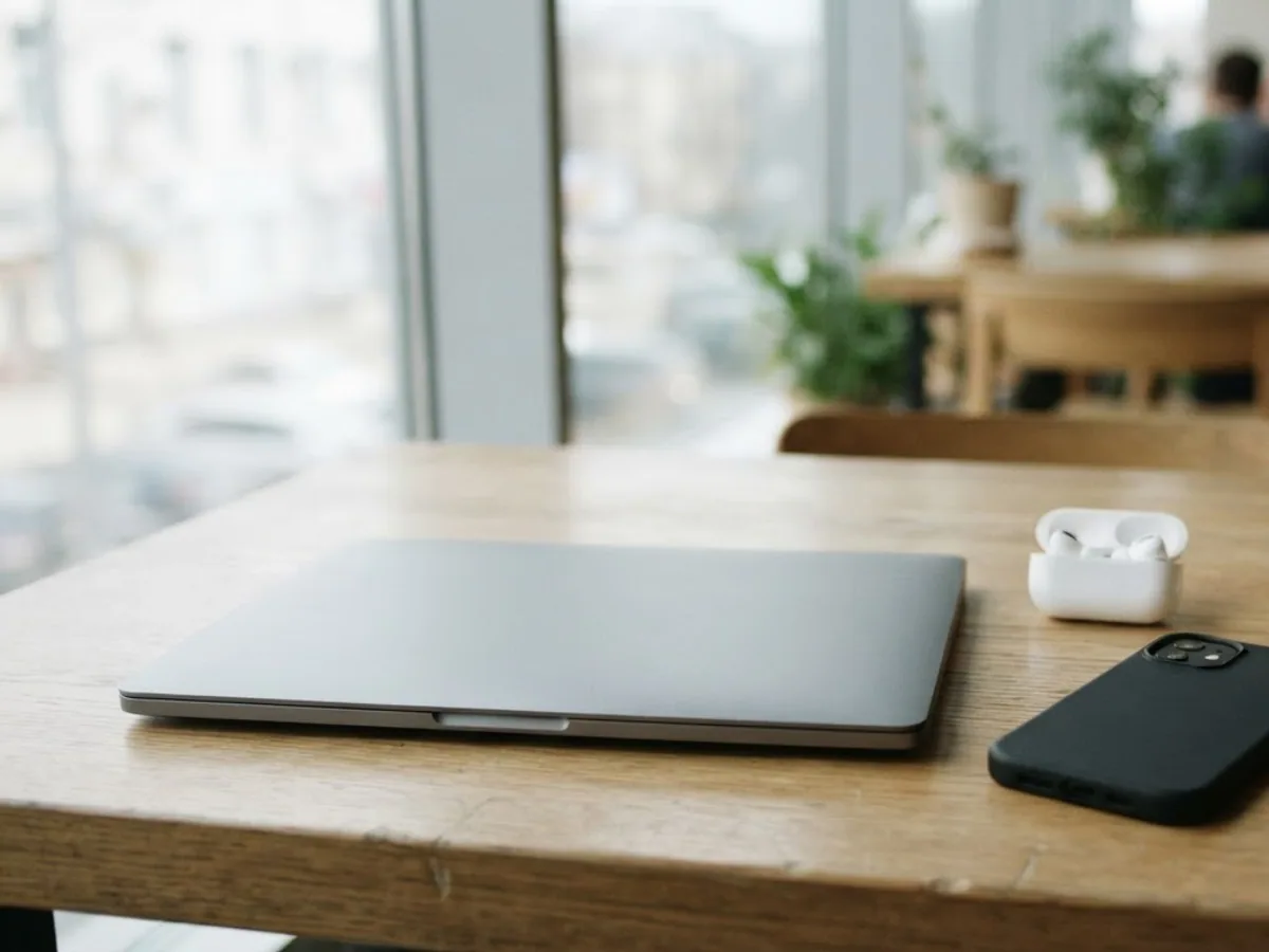 Closed laptop, open earbuds case, face-down phone and a coffee cup on a shared desk