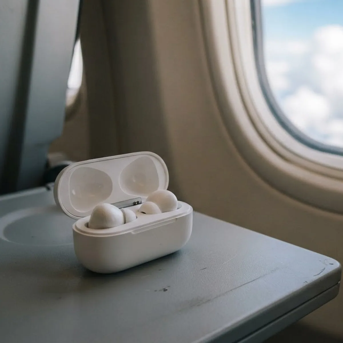 Open white earbuds case with earbuds inside on an airplane tray table beside the window
