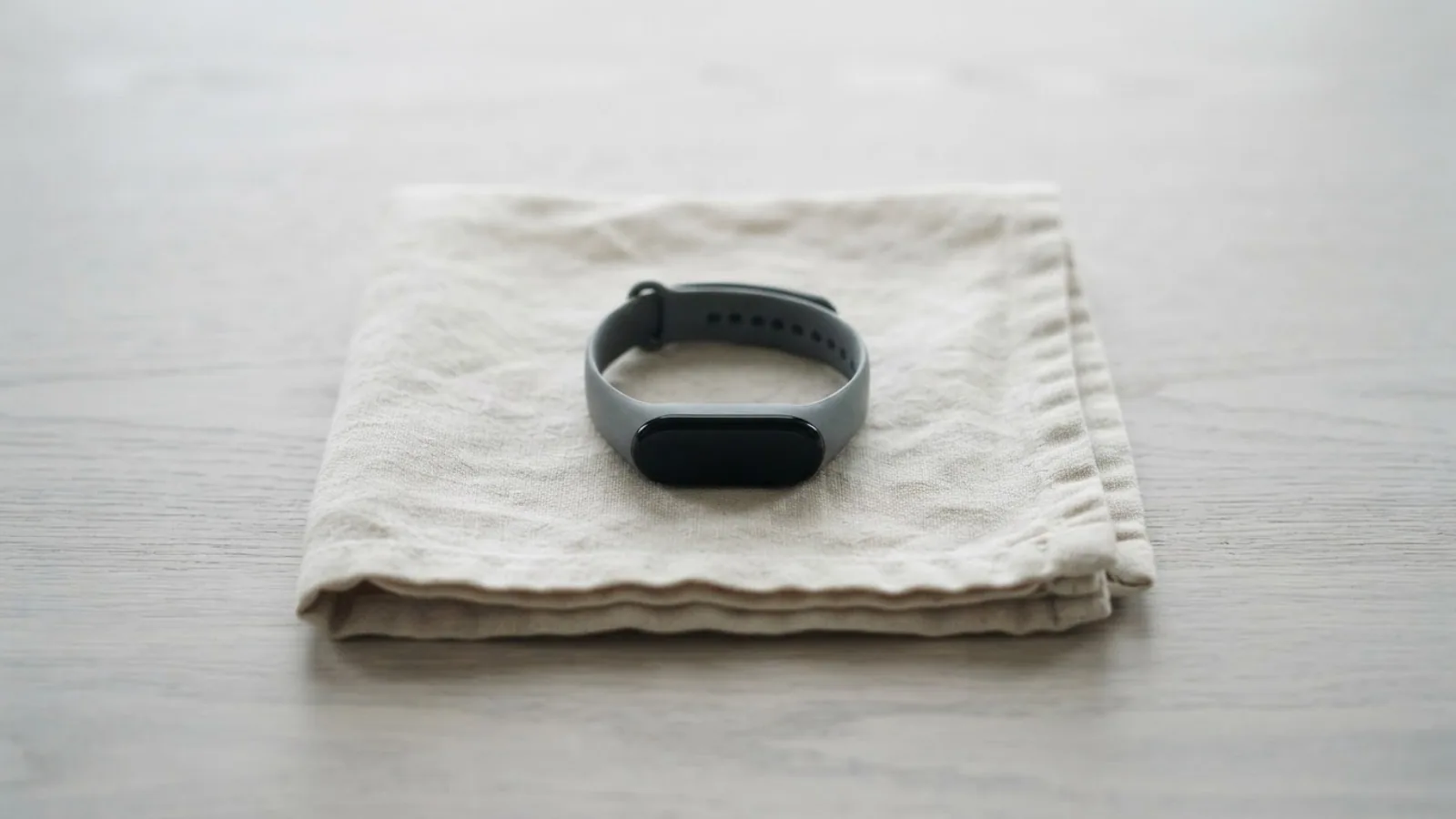 A slim fitness tracker with a grey band resting on a folded linen cloth on a pale wooden table