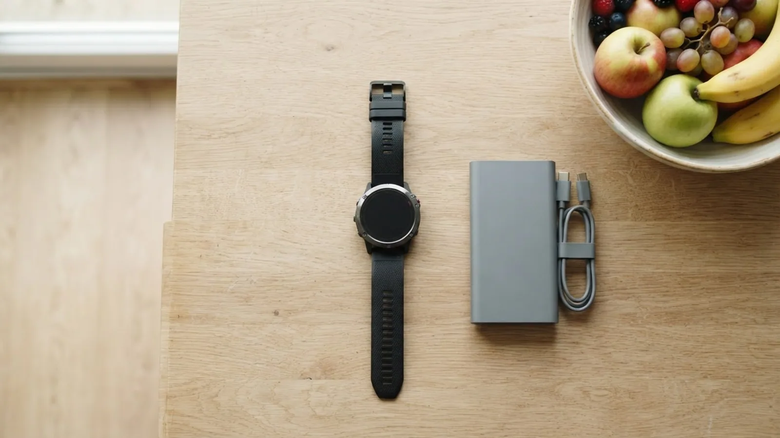 Top-down view of a round sports watch beside a grey power bank and a coiled cable on a kitchen table, with a fruit bowl