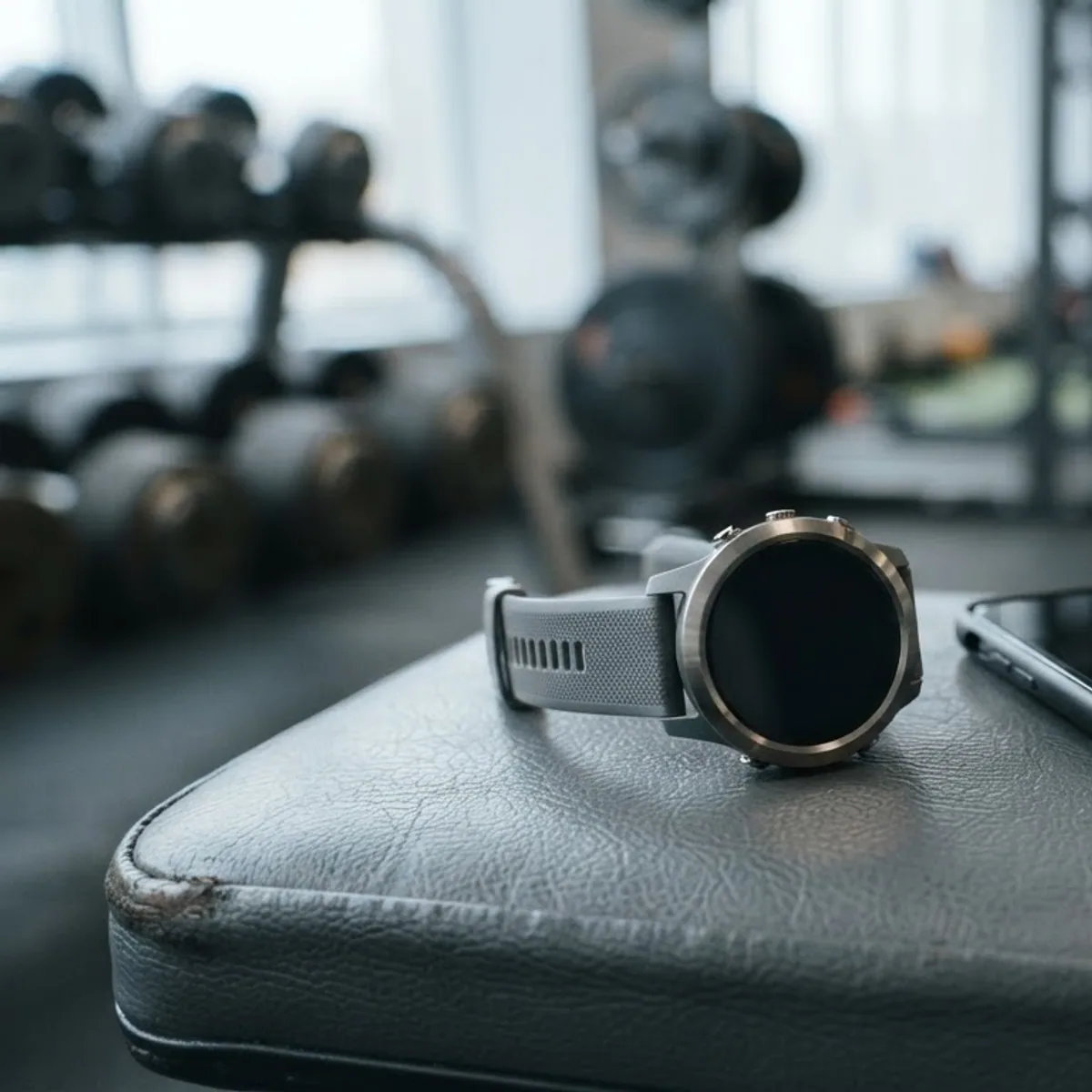 A round sports watch with a grey strap next to a phone on a padded gym bench, with weights blurred behind
