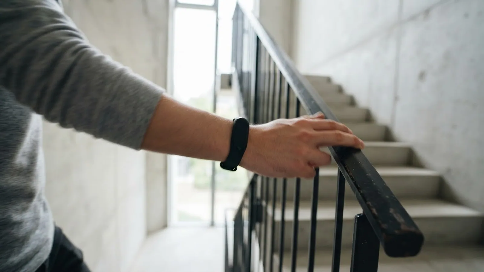 A hand on a stair handrail while climbing, with a slim fitness tracker band on the wrist, in a bright stairwell.