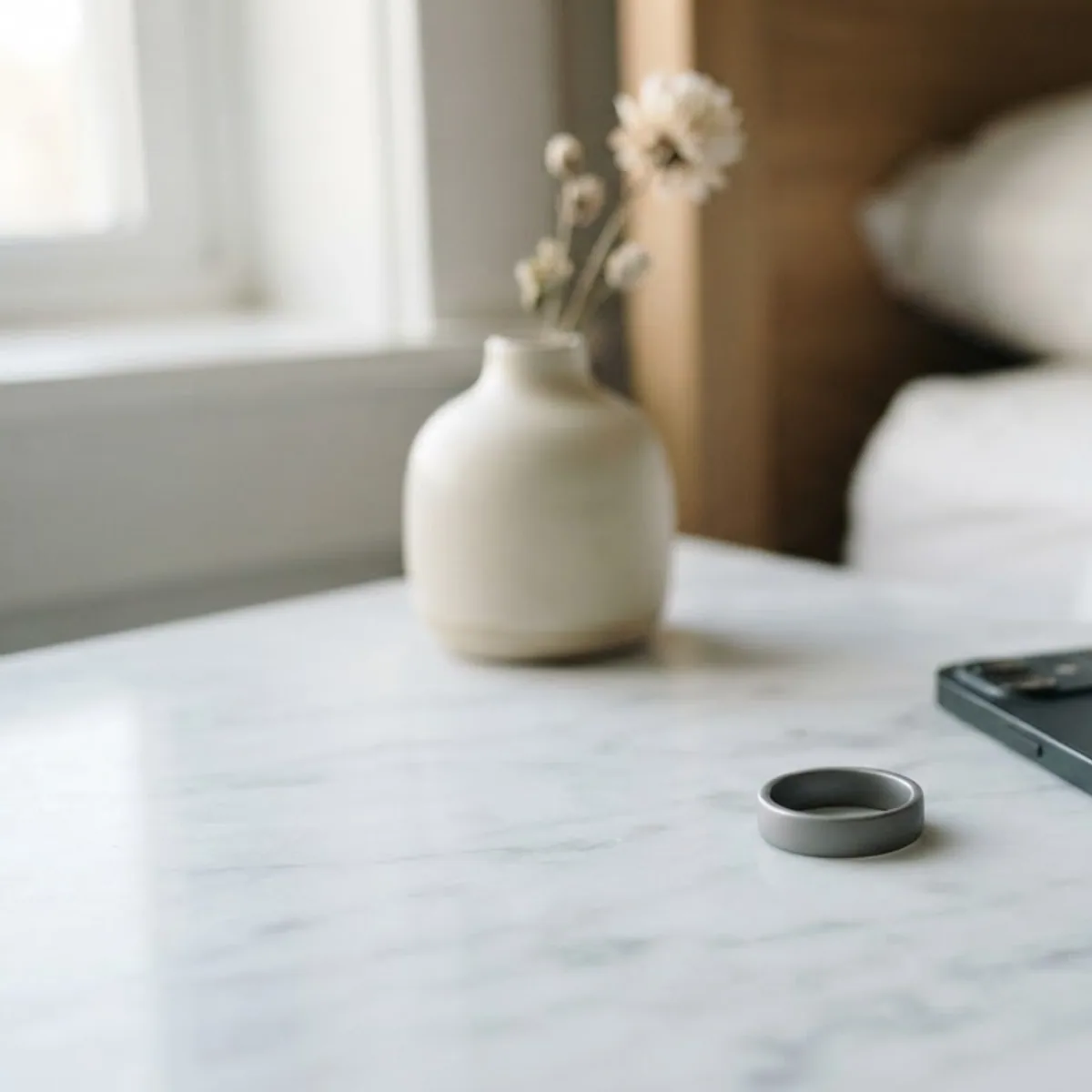 Plain grey smart ring on a white marble bedside table beside a smartphone lying face down