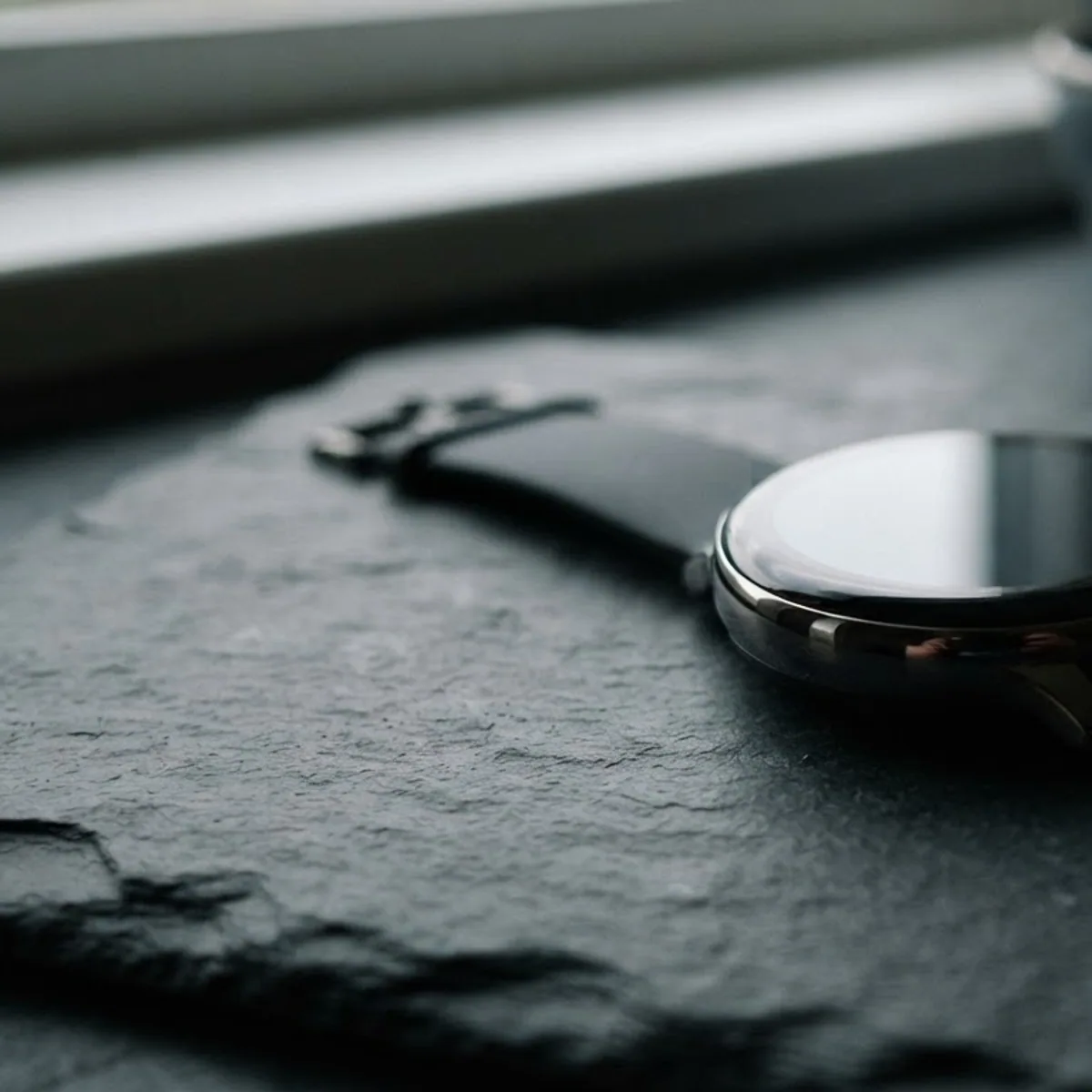 A round smartwatch with a completely black screen lying on a dark slate surface in low side light.