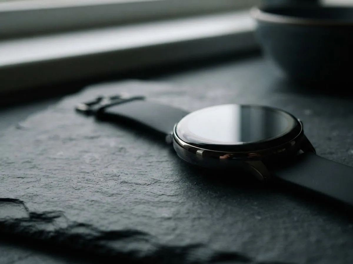 A round smartwatch with a completely black screen lying on a dark slate surface in low side light.