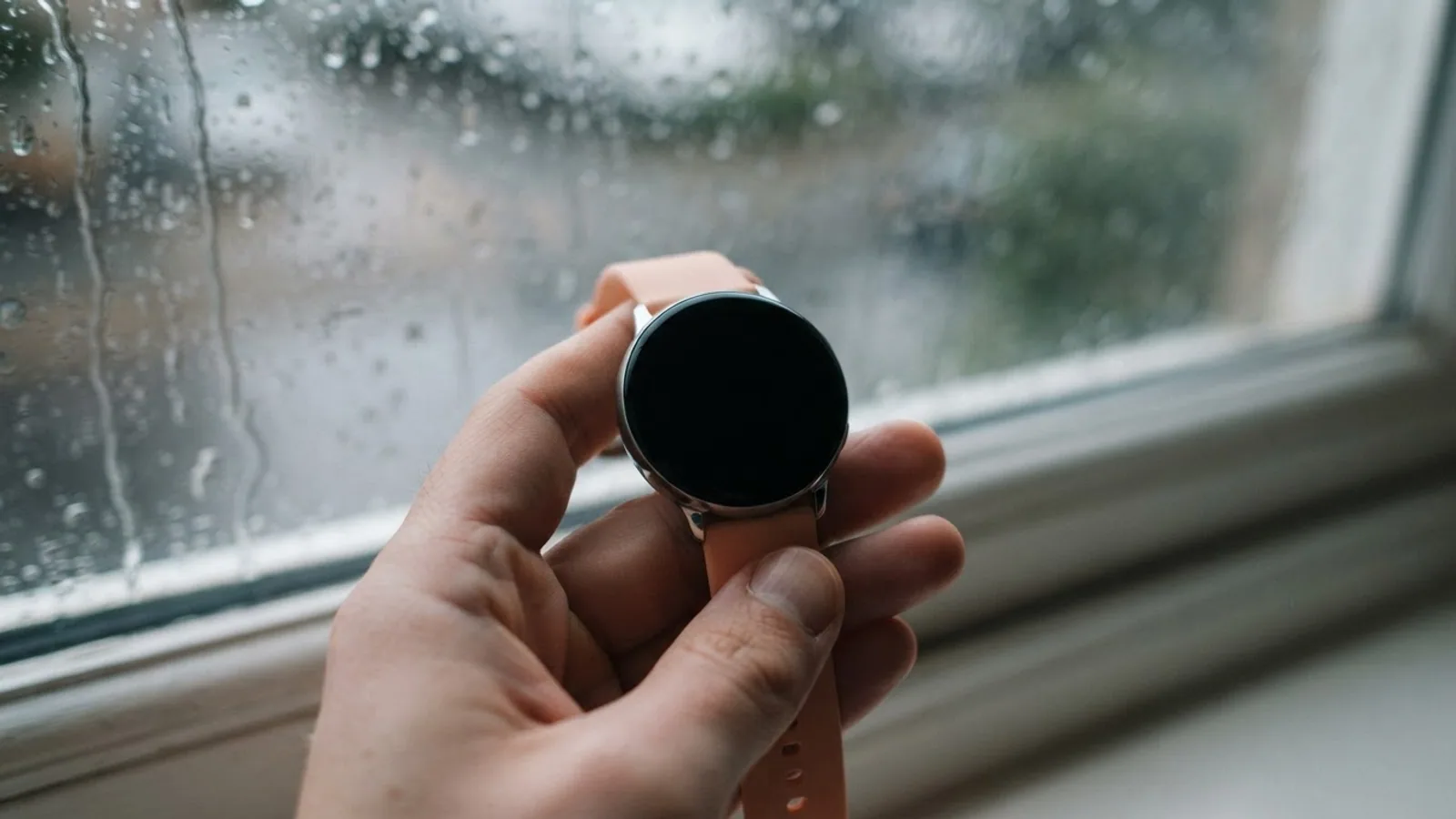 A hand holding a round smartwatch with a black screen beside a rainy window.