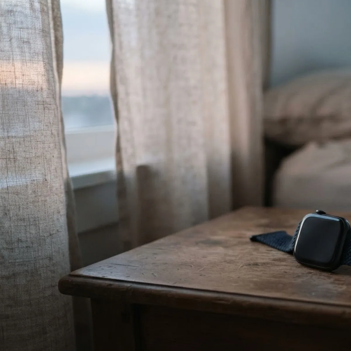 A square smartwatch with a braided band on a bedside table next to a glass of water at dawn