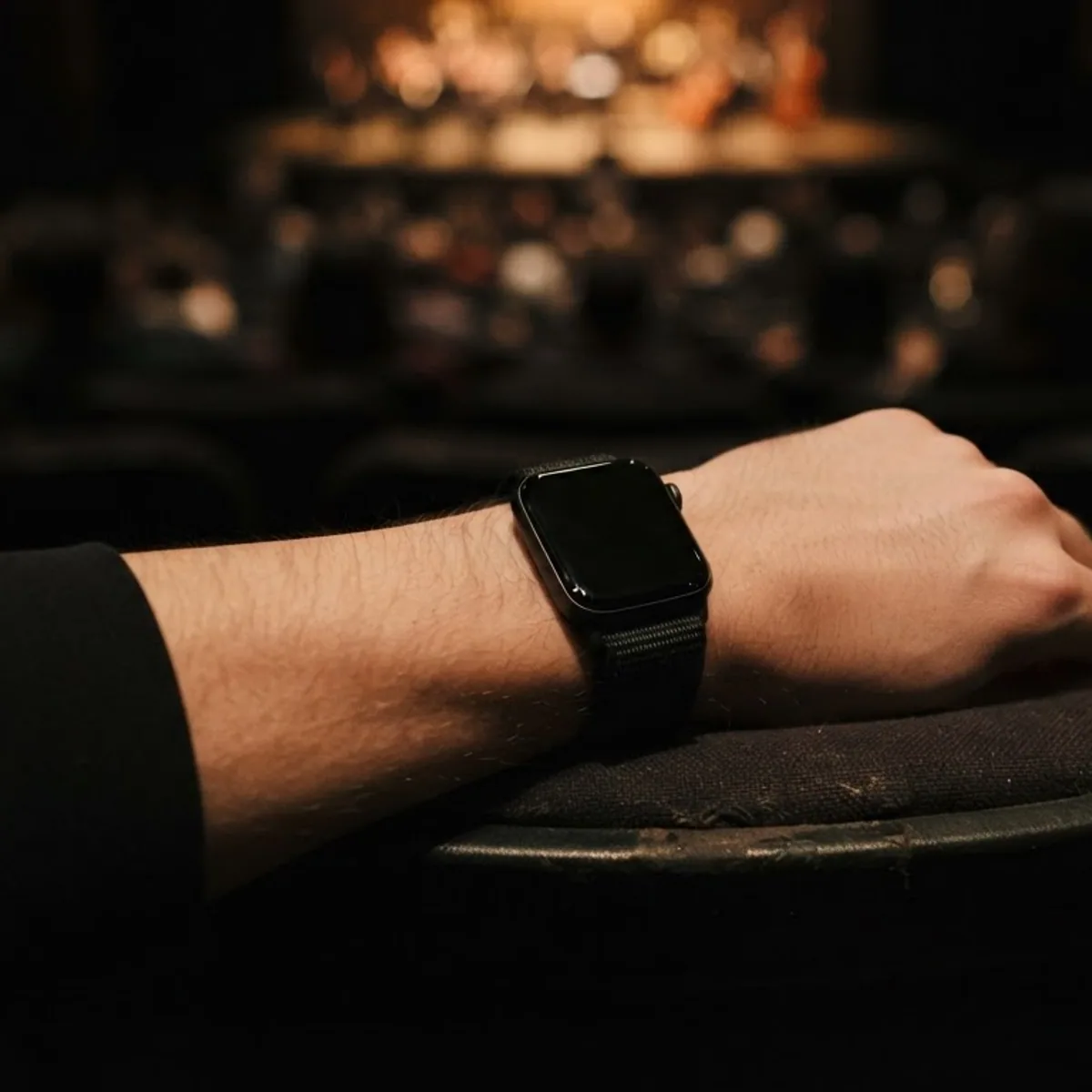 A forearm wearing a square smartwatch resting on a theater seat armrest in a dim auditorium