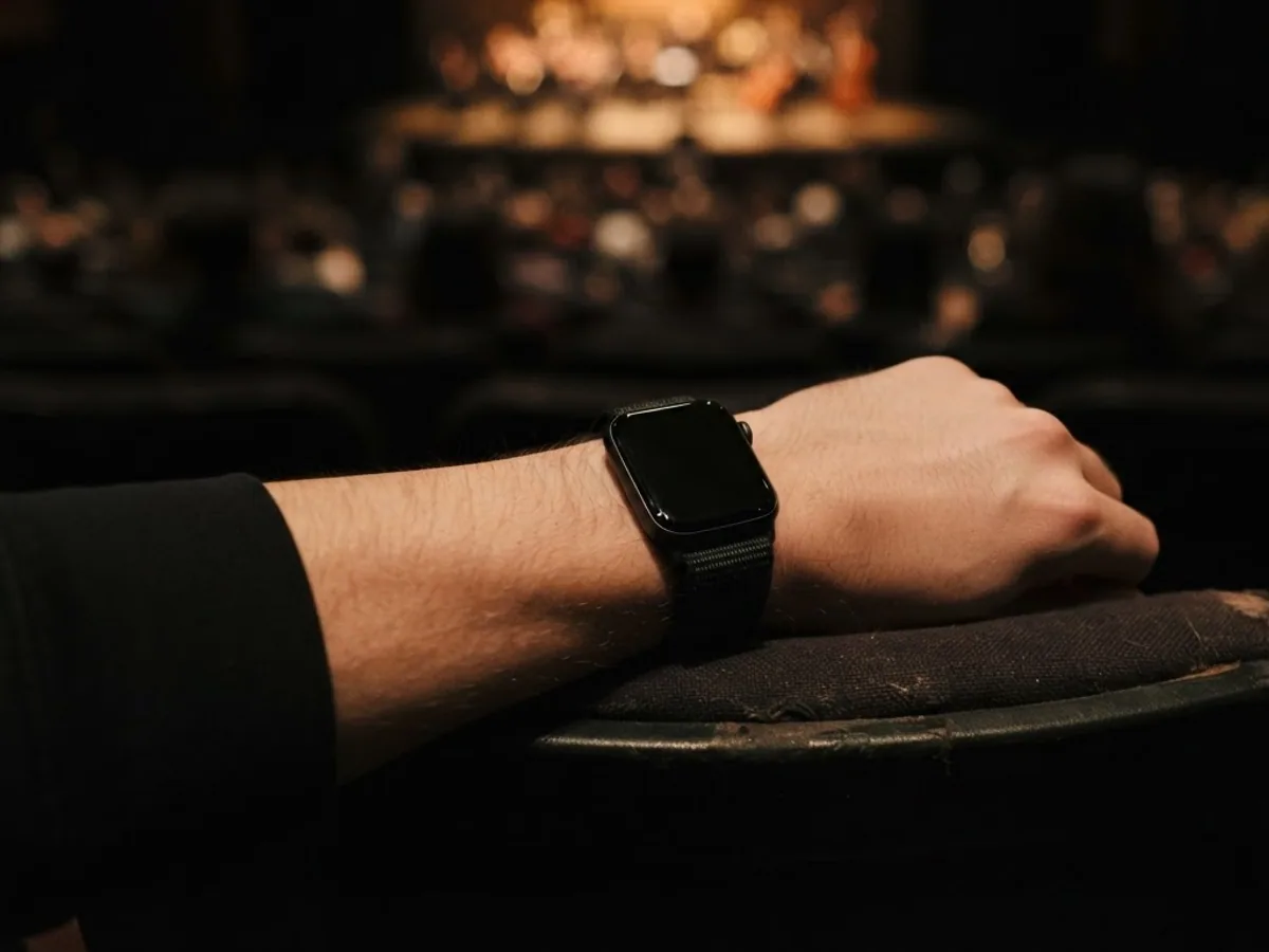 A forearm wearing a square smartwatch resting on a theater seat armrest in a dim auditorium