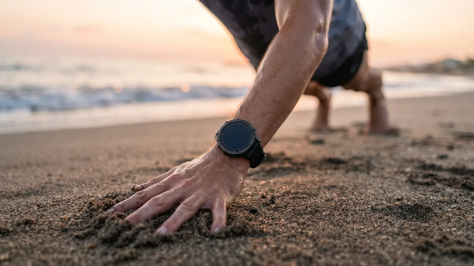 A wrist wearing a round sports watch as the hand presses into the sand while stretching on a beach at sunrise