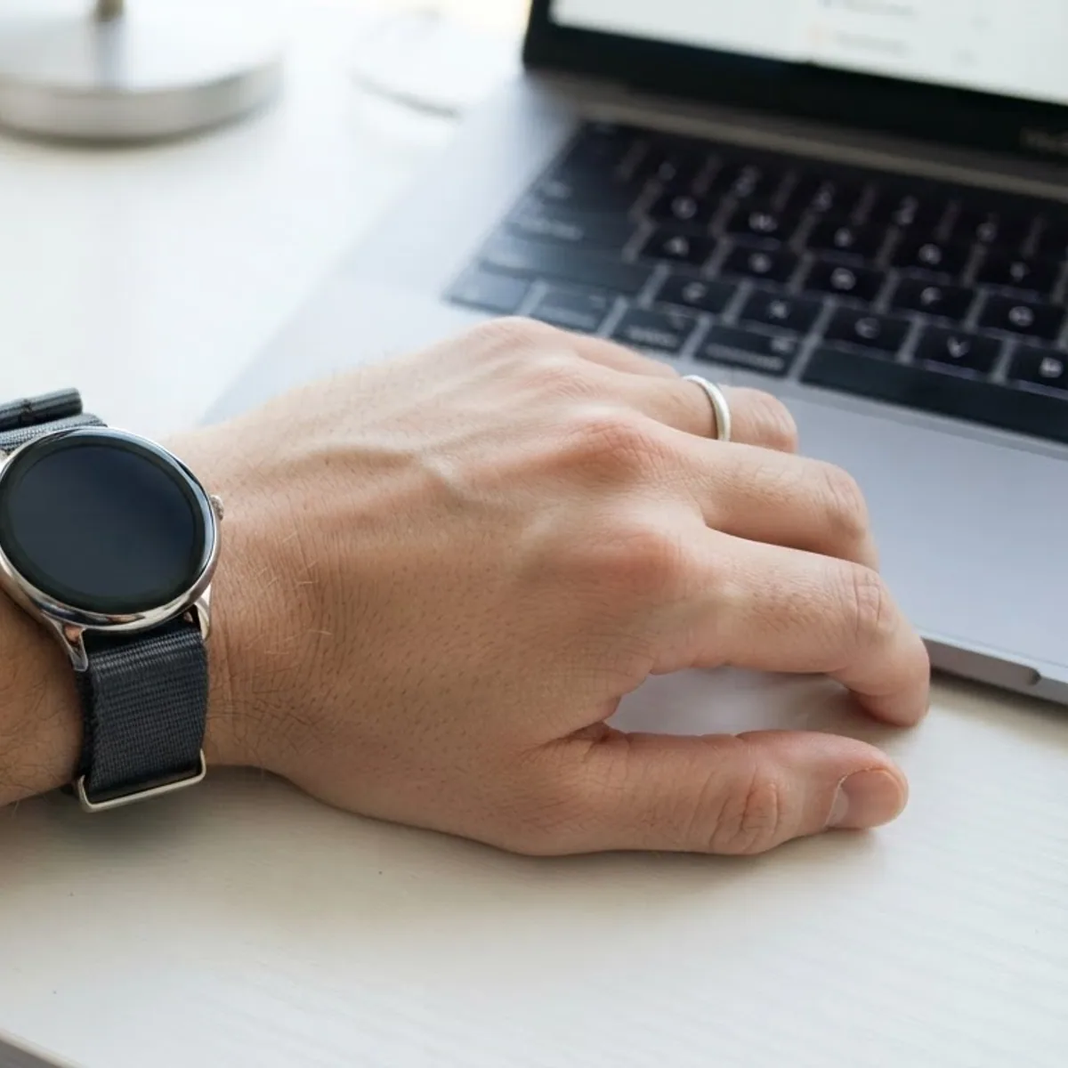 A wrist wearing a slim round smartwatch with a fabric band, resting beside a laptop keyboard